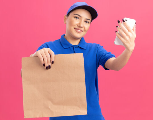 拿着身着蓝色制服 戴着帽子 手持纸包 站在粉色墙壁上 面带微笑的快乐年轻送货员包装欢呼纸