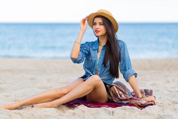 海滩美丽的女人 棕褐色的身体 丰满的红唇和长长的腿 在热带阳光明媚的海滩上摆姿势穿着短上衣 短裤和草帽乐趣坐着欢乐