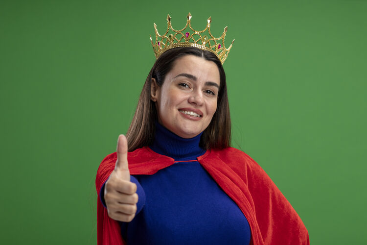 表演带着微笑的年轻超级英雄女孩戴着王冠 在绿色背景上孤立地竖起大拇指穿皇冠超级英雄