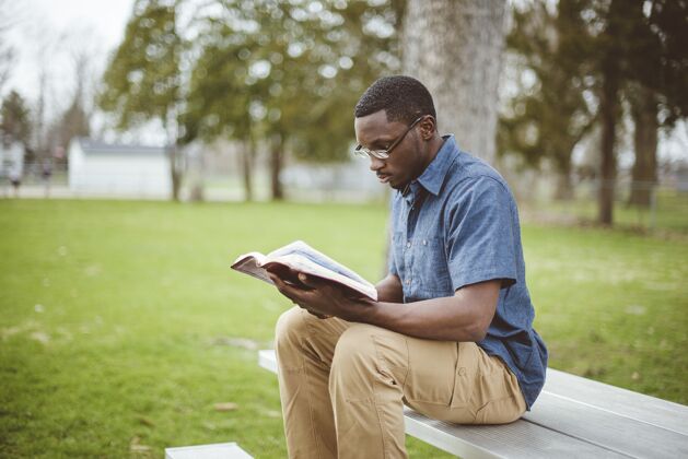 徒年轻的非洲裔男性坐在长椅上读圣经公园男性长凳