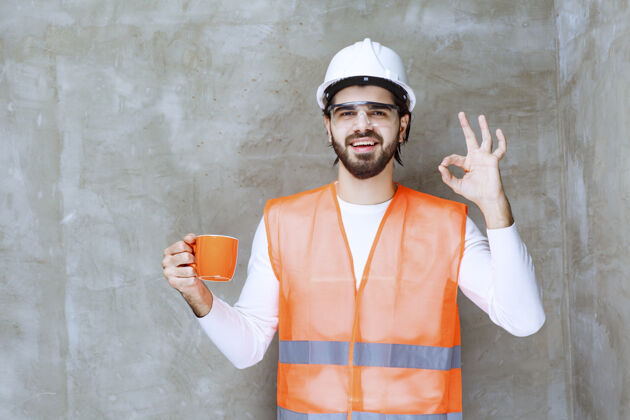 聪明戴着白色头盔的工程师拿着一个橙色的杯子 享受着饮料的味道人体模特提升人