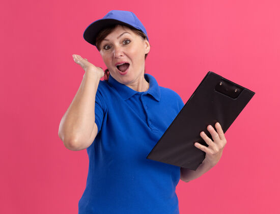 女人身穿蓝色制服 戴着帽子的中年送货妇女站在粉红色的墙上 高兴而惊讶地看着前面的剪贴板快乐举行剪贴板