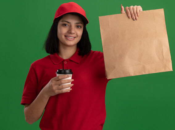 微笑身穿红色制服 头戴鸭舌帽的年轻送货小姐 手拿纸包和纸杯 脸上带着微笑站在绿色的墙上帽子交货持有