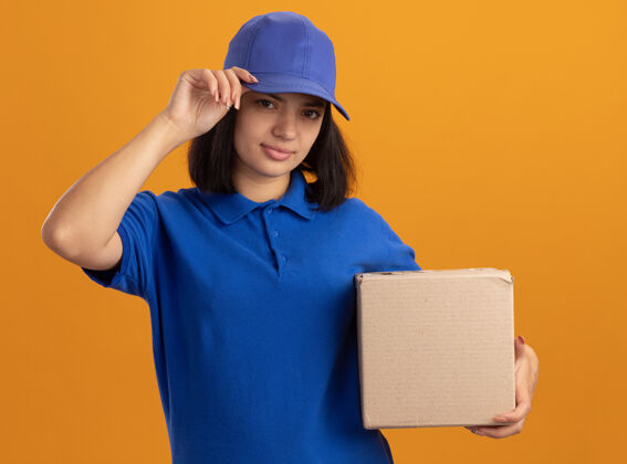 立场身着蓝色制服 戴着帽子的年轻送货员站在橙色的墙上 脸上带着自信的微笑 手里拿着纸箱交货脸信心