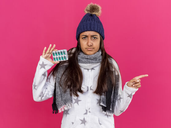 年轻未出租的年轻生病的女孩戴着冬天的帽子与围巾举行药片和点在一边孤立的粉红色背景与复制空间围巾点女孩