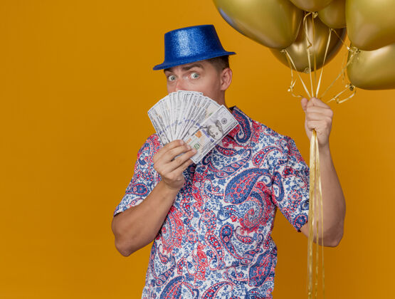 年轻人惊讶的年轻人戴着蓝色帽子拿着气球 脸上贴着现金 背景是橙色 还有复印空间小伙子帽子抱着