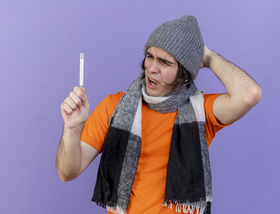 手一个戴着冬天帽子 戴着围巾 手放在头上看温度计的年轻病患 被隔离在紫色的背景上帽子体温计年轻人