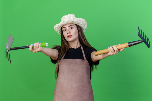锄头印象深刻的美丽的园丁女孩穿着制服戴着园艺帽子拿着耙子锄头耙在相机上孤立的绿色背景穿耙子制服