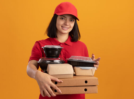 包装身穿蓝色制服 头戴鸭舌帽 手持比萨饼盒和食品包的年轻送货女孩站在橙色的墙上 微笑着 快乐而积极送货正面微笑