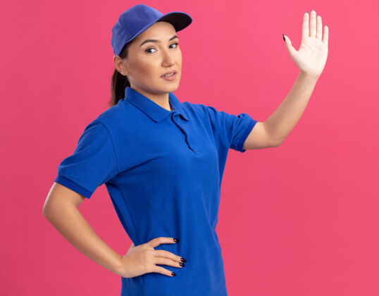 开放身穿蓝色制服 头戴鸭舌帽的年轻女送货员微笑着自信地举起手站在粉色的墙上微笑帽子年轻