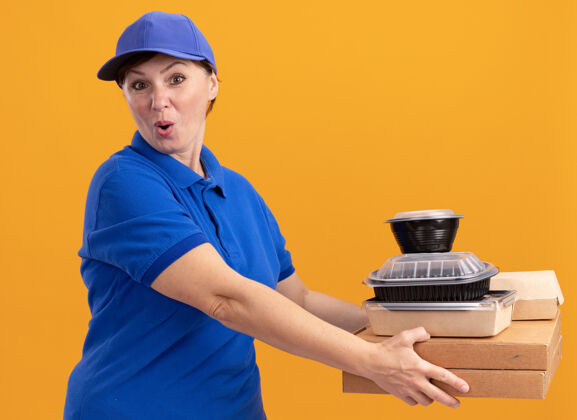 帽子身穿蓝色制服 头戴鸭舌帽的中年女送货员拿着披萨盒和食品包 站在橘色的墙上 高兴而又惊讶地看着前面惊喜送货盒子