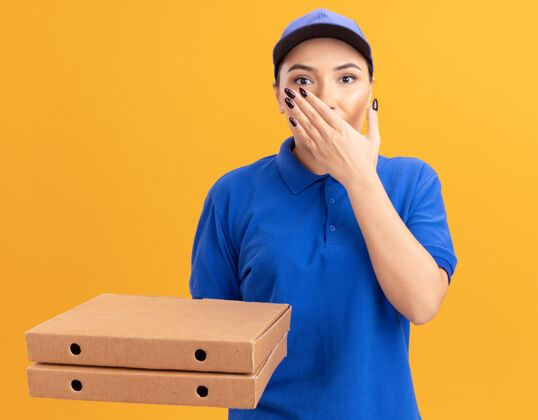 披萨身穿蓝色制服 戴着帽子的年轻送货员拿着披萨盒看着前面 震惊地用手捂住嘴巴站在橙色的墙上制服站着送货