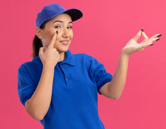 制服穿着蓝色制服 戴着帽子的年轻送货员用食指指着眼睛 用手指站在粉红色的墙上做着赚钱的手势眼睛手指制造