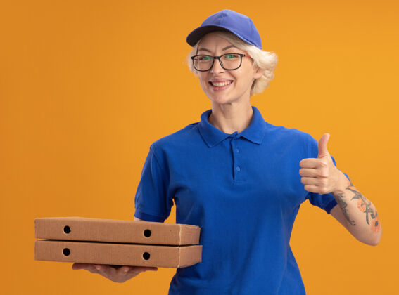 展示身穿蓝色制服 戴着眼镜 举着披萨盒 竖起大拇指 微笑着站在橙色墙上的年轻送货员披萨眼镜拿着