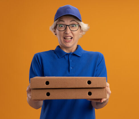 兴奋身穿蓝色制服 戴着眼镜 拿着比萨饼盒的年轻送货员在橙色的墙上兴奋而快乐女人盒子送货