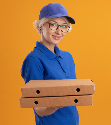 戴着身穿蓝色制服 戴着眼镜 拿着比萨饼盒的年轻女送货员望着橙色墙上自信微笑的摄影师女人帽子眼镜