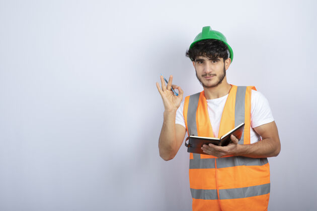 工作帅气的男工程师戴着绿色的安全帽 笔记本立在白色的墙上高质量的照片建筑师建筑笔记本