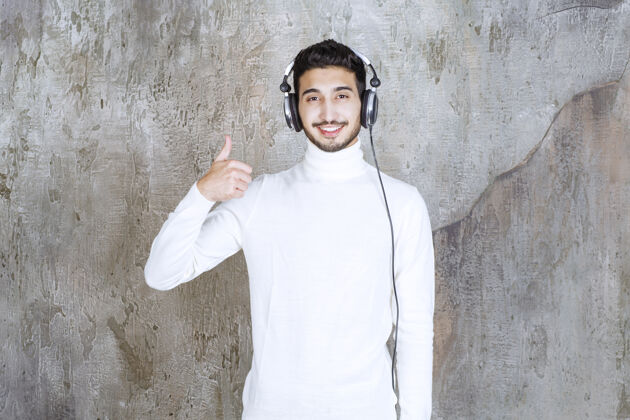 享受穿白毛衣的男人戴着耳机 听着音乐 表现出积极的手势成人满意好