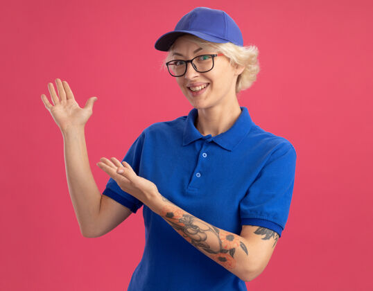 制服身着蓝色制服 头戴帽子的快乐女快递员微笑着 双手搭在粉红色的墙上 愉快地展示复印空间介绍年轻微笑