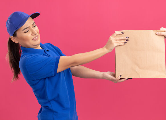 纸身穿蓝色制服 头戴鸭舌帽 表情厌恶的年轻女送货员站在粉红色的墙上收包裹递送女人制服