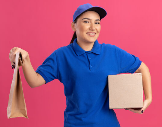 站着身穿蓝色制服 头戴鸭舌帽的年轻女送货员拿着纸包和纸板箱望着前方 微笑着自信地站在粉色的墙上纸板自信抱着