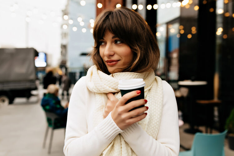 咨询外面的画像是迷人的漂亮女士 棕色头发 穿着白色毛衣和围巾 在灯光下的街上喝咖啡魅力搜索学生