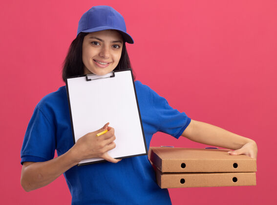盒子身穿蓝色制服 头戴鸭舌帽 拿着披萨盒和空白页的剪贴板等待签名的年轻送货女孩站在粉色的墙上空白站着剪贴板
