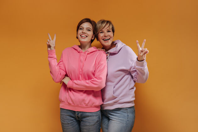 橙色背景时尚的两位女士 短发 亮粉色宽边连帽衫 酷酷牛仔裤 在橙色背景上微笑着展示和平标志年轻中等成熟