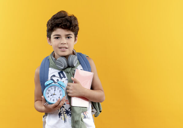 小学生高兴的小男孩戴着书包和耳机拿着一本书与闹钟隔离在黄色背景与复印空间小耳机请戴