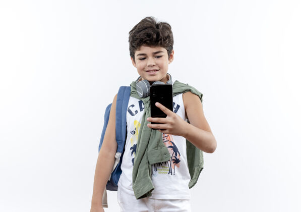 耳机高兴的小男生背着书包 戴着耳机 拿着手机 看着孤立的白色背景背小学生抱着