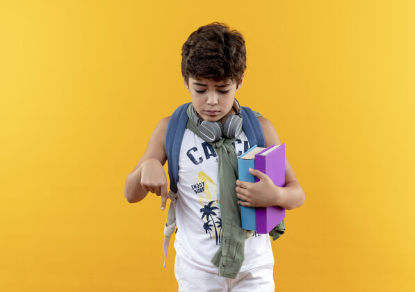 伤心看着那个戴着背包和耳机拿着书的悲伤的小男孩 指着黄色背景上孤立的 有复印空间的下来学校点男孩
