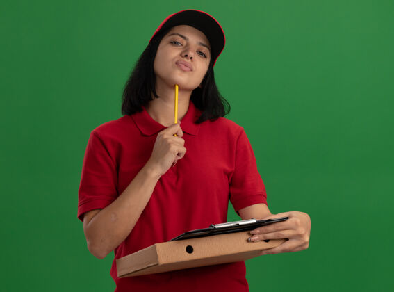 制服身穿红色制服 头戴鸭舌帽 拿着披萨盒和铅笔的剪贴板的年轻送货女孩站在绿色的墙上立场送货举行