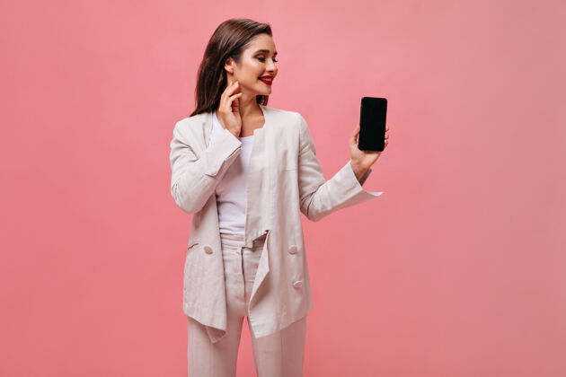 站立心情好的女士拿着粉色背景的智能手机穿着米色办公服的可爱女商人在孤立的背景上看电话粉色夹克肖像