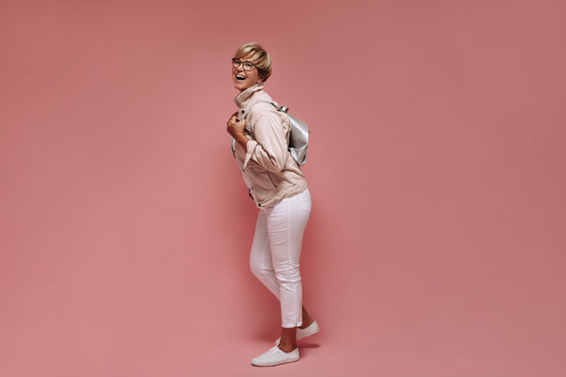 肖像一张短发戴眼镜 穿着紧身轻便裤 白色运动鞋和夹克的女士在粉色背景下笑着拿着包的全貌照片室内女性夹克