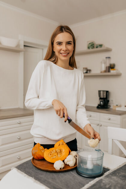 失落穿着白衬衫的快乐兴奋的年轻女士一边在厨房里煮蔬菜一边对着镜头摆姿势金发爱好阅读