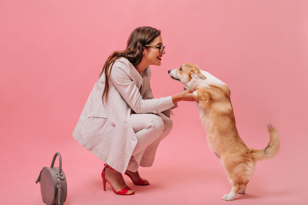 肖像穿米色西装的女人和粉色背景的狗玩戴着眼镜和红色高跟鞋的可爱漂亮女孩看着柯基犬微笑着卷发站立白色