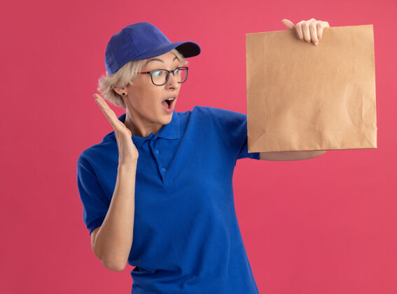 惊人身穿蓝色制服 头戴鸭舌帽的年轻女送货员拿着纸包 看着粉色的墙上 惊讶又惊奇举行制服包装