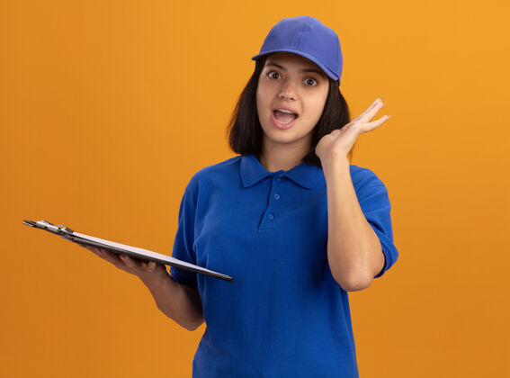制服身穿蓝色制服 戴着帽子的年轻送货女孩站在橙色的墙上 微笑着惊讶地站着剪贴板举行惊喜