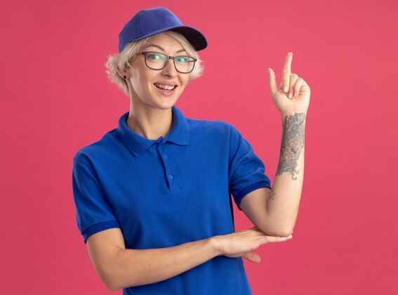 女人身着蓝色制服 头戴鸭舌帽的快乐年轻女送货员微笑自信地伸出食指站在粉红色的墙上年轻帽子信心