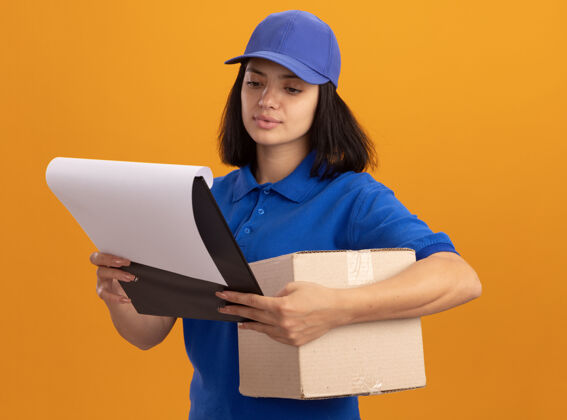 严重穿着蓝色制服 戴着帽子的年轻送货女孩拿着纸板箱和空白页的剪贴板 严肃地看着站在橙色墙上的页面立场脸交货