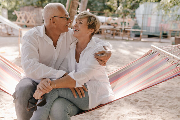 肖像快乐的老妇人 金发碧眼 穿着衬衫和牛仔裤 坐在格子吊床上 在沙滩上和微笑的白发男人拥抱祖父母海滩拥抱
