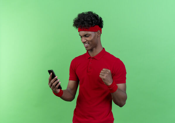 非裔美国人快乐的年轻黑人美国运动男子戴着头带和腕带举行 并看着电话 并显示是的手势孤立的绿色背景欢乐手势头带