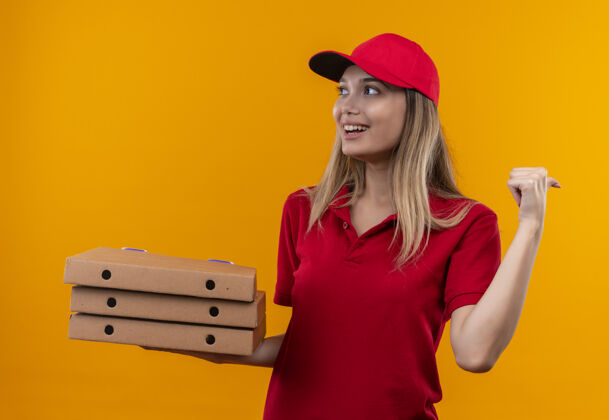 制服看着面带微笑的年轻送货女孩穿着红色制服 戴着帽子拿着披萨盒 指着橙色背景上孤立的一面壁板年轻人穿