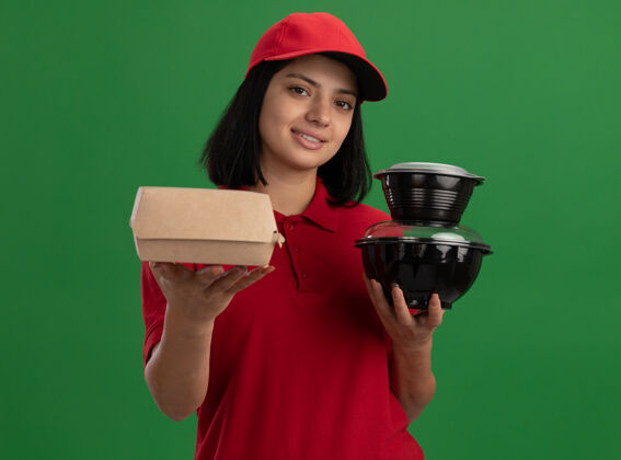 帽子身着红色制服 头戴鸭舌帽 手持食品包装 面带微笑 友好地站在绿色墙壁上的快乐少女盒子朋友女孩