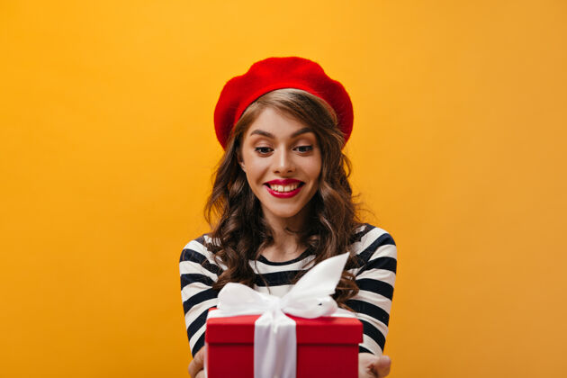 女孩身着红色贝雷帽和条纹衬衫的女士手持礼品盒迷人的女士 波浪式发型 戴着明亮的帽子 穿着时髦的衬衫摆姿势肖像头发成人