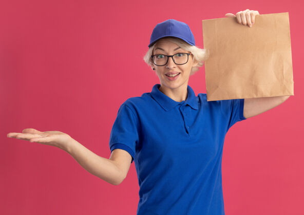 介绍身穿蓝色制服 戴着眼镜 手举纸包 手举粉色墙壁 展示复印空间的年轻送货妇女眼镜包装女人
