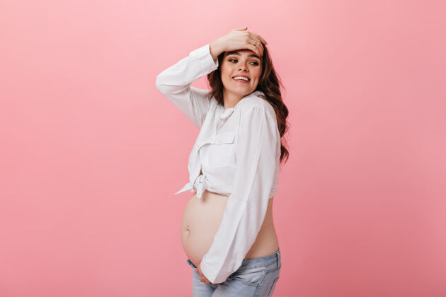 微笑情绪化快乐的孕妇穿着白色短衬衫和牛仔裤 在孤立的粉色背景下抚摸头发和微笑怀孕肚子粉色