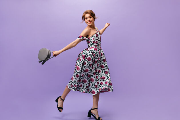 花穿着花裙的年轻女子拿着手提包跳了起来迷人可爱的年轻女士 时尚的发型 在夏季的服装摆姿势微笑紫色肖像