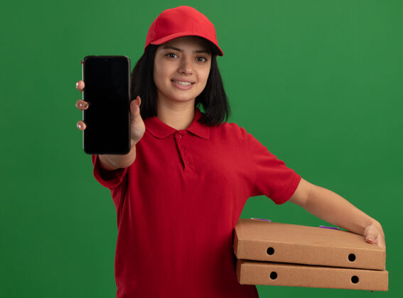 表演身穿红色制服 头戴鸭舌帽 手持披萨盒 脸上挂着微笑的智能手机的年轻送货女孩站在绿色的墙上拿着递送盒子