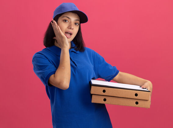 披萨身穿蓝色制服 头戴鸭舌帽 拿着比萨饼盒和剪贴板的年轻送货女孩站在粉红色的墙上 惊讶不已女孩剪贴板制服